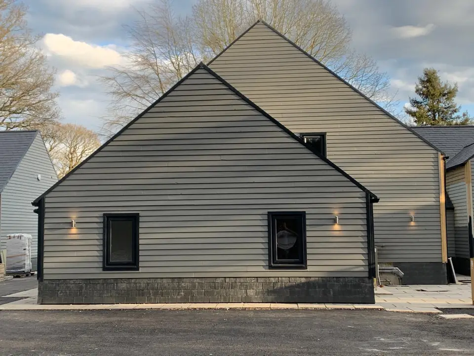 Gable end of a barn building in grey horizontal cladding above a dark plinth, with wall lights either side of the windows