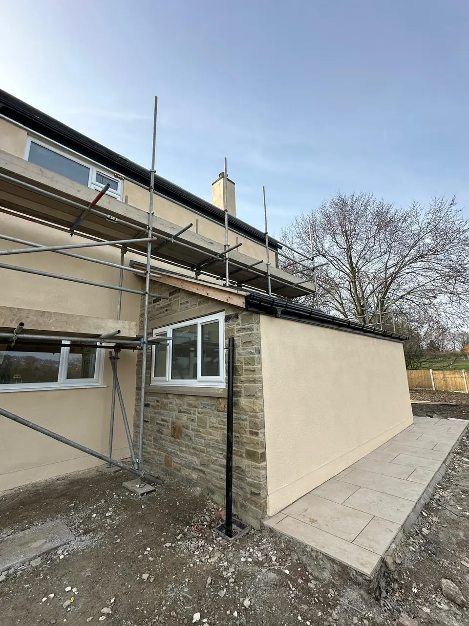 Cream rendered extension wall meeting the stone house, with scaffolding still up along the main house