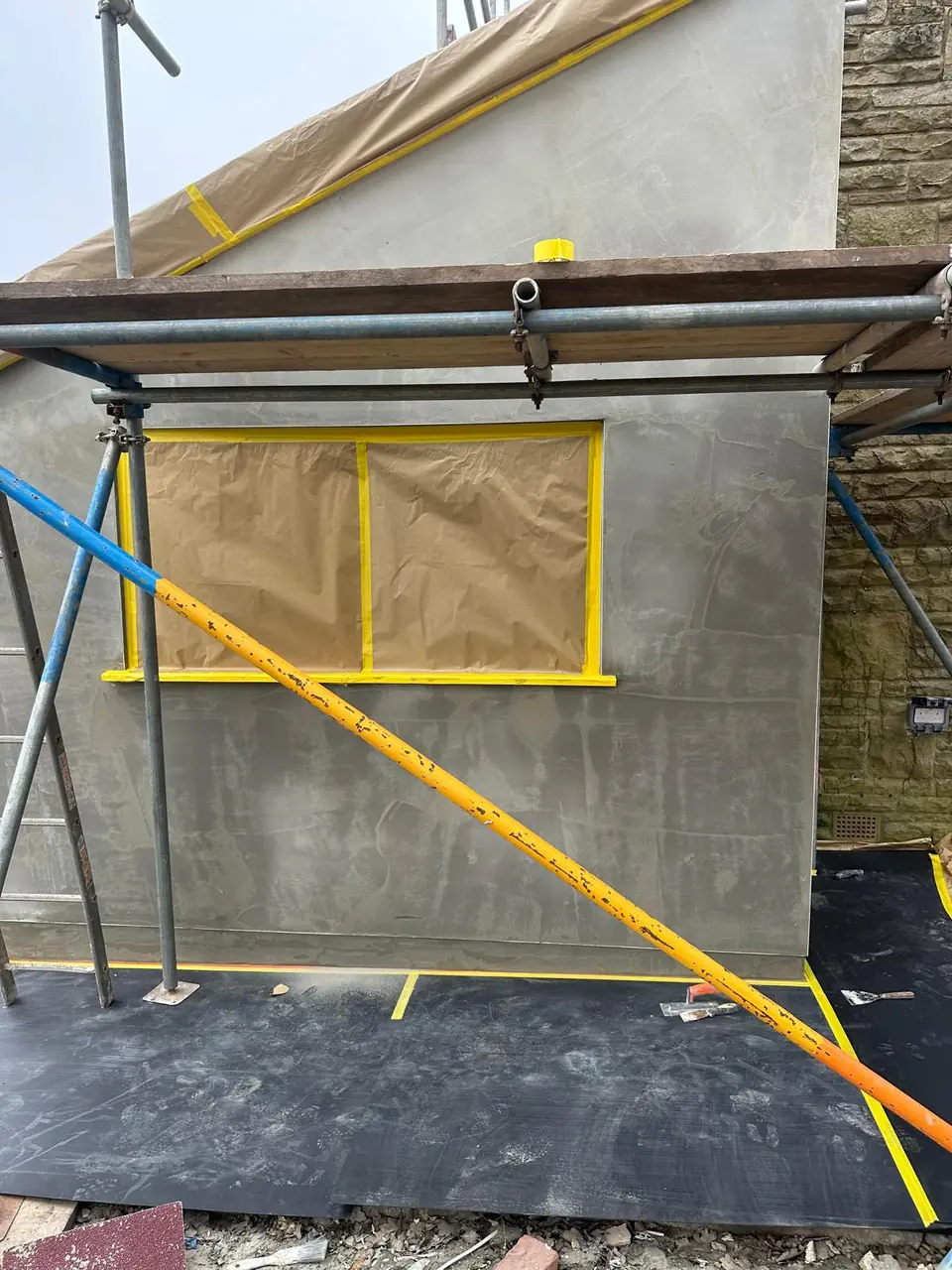 Extension wall in a grey base coat, with the window masked off in yellow tape before the finish coat